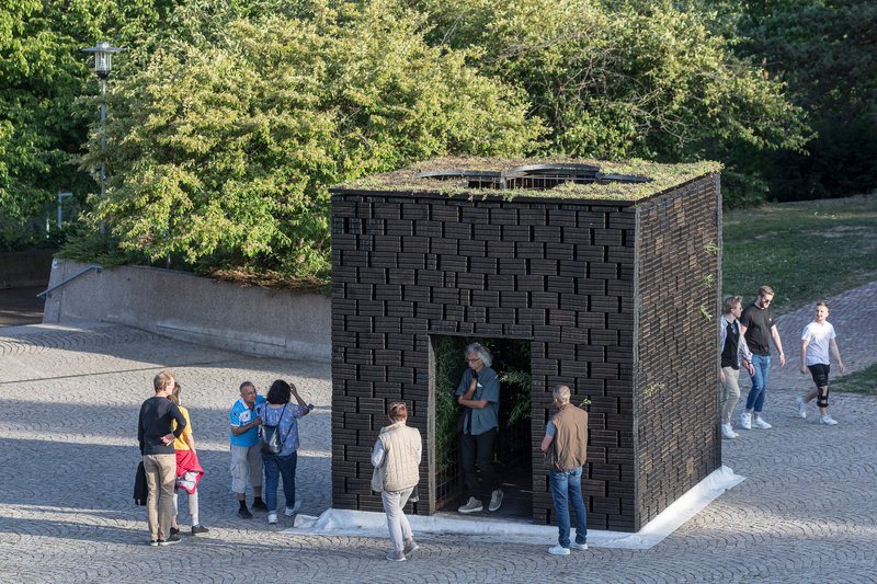 Dark woven brick pavilion with green roof and arched opening surrounded by visitors in afternoon light