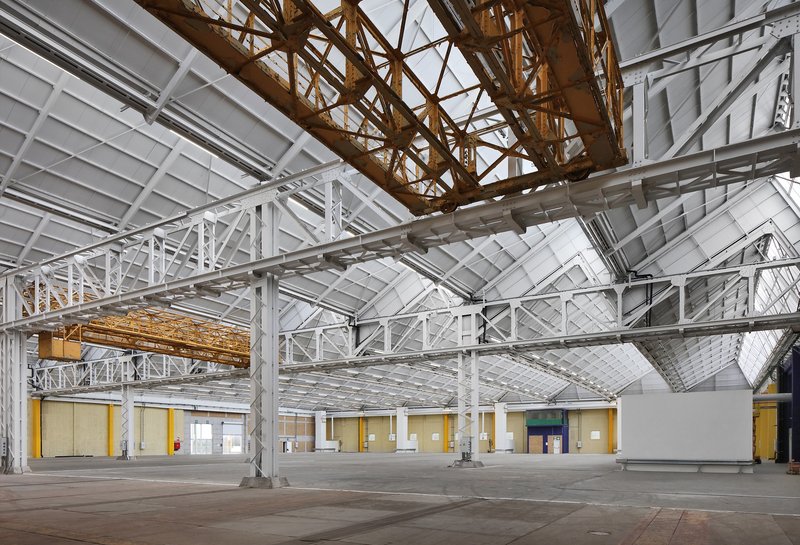 Interior hall with exposed white and gold steel trusses supporting a sawtooth ceiling above yellow-doored volumes