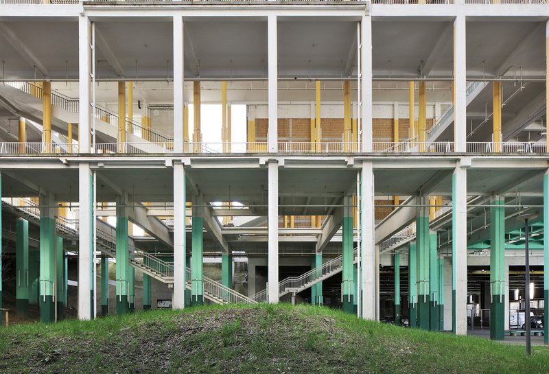 Multilevel concrete frame with yellow and green columns visible beneath white balustrades and sloped grassy embankment