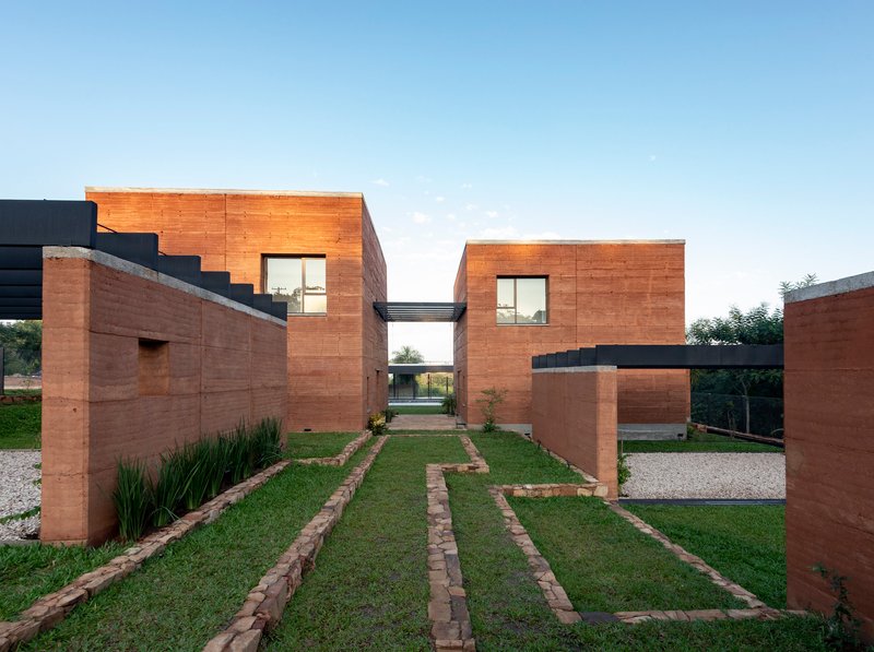 Front courtyard with rammed earth walls, planted grass strips and gravel paths leading to a skybridge