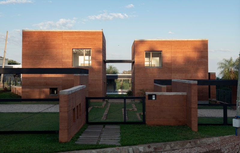 Street view of paired rammed earth volumes connected by a bridge under evening light