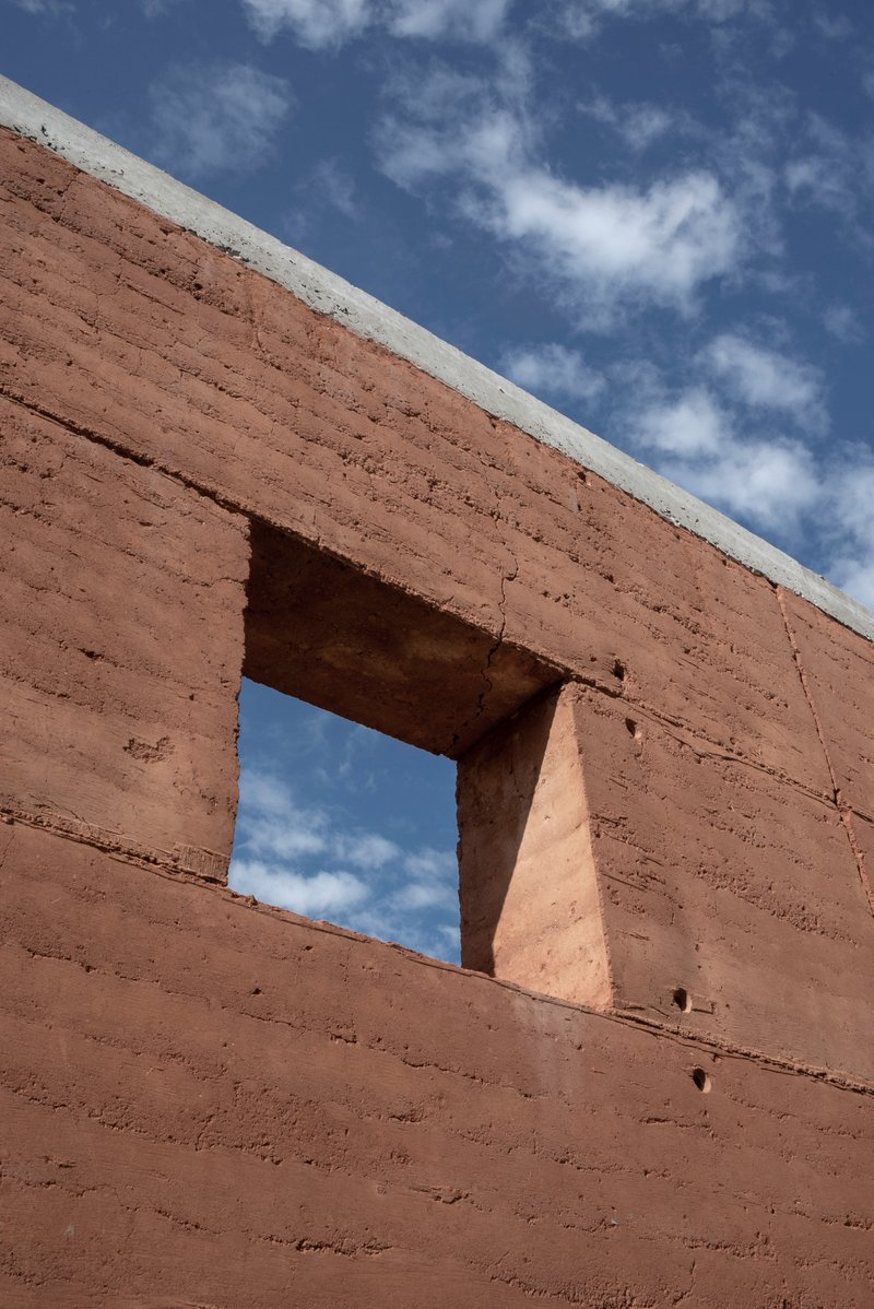 Rammed earth wall with rectangular window opening framing blue sky and white clouds