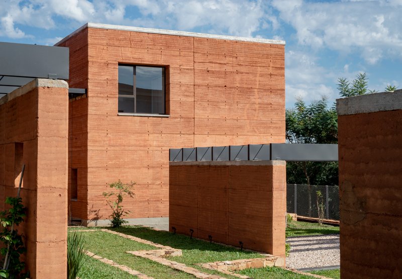 Two-storey rammed earth volume with metal bridge connection under a cloudy afternoon sky