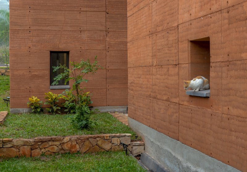 Rammed earth facade with recessed window and planted bed beside a stone garden sculpture on a shelf