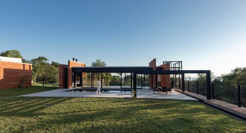 Glass pavilion with steel frame connecting rammed earth volumes across a sloped lawn at dusk