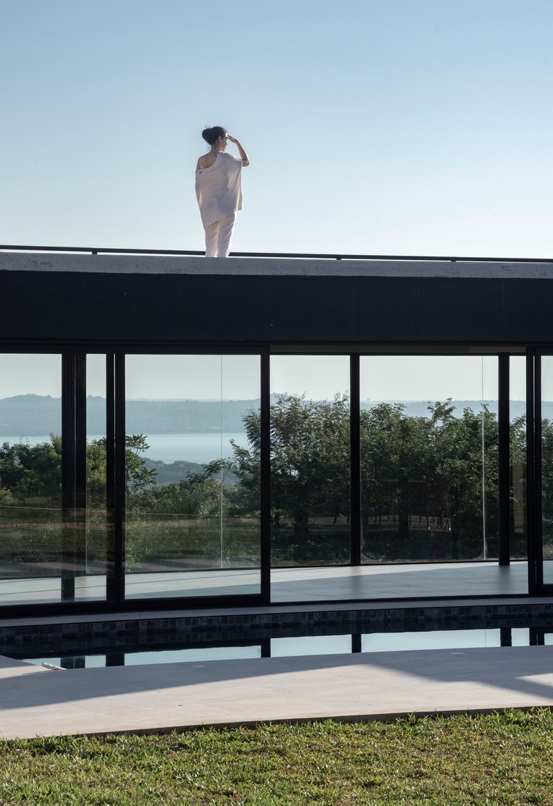 Figure standing on the flat roof above the glass-walled pool pavilion overlooking distant hills