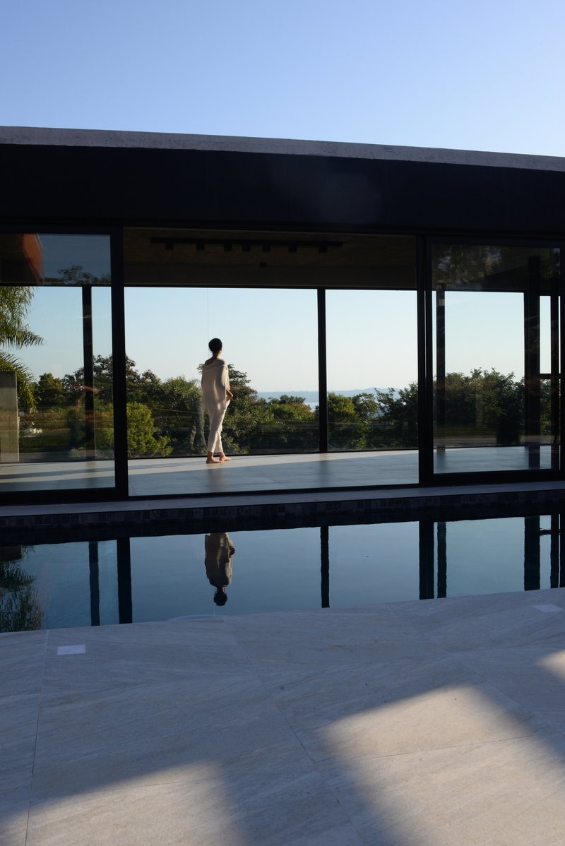 Pool terrace with figure reflected in still water beside the steel-framed glass pavilion at sunset