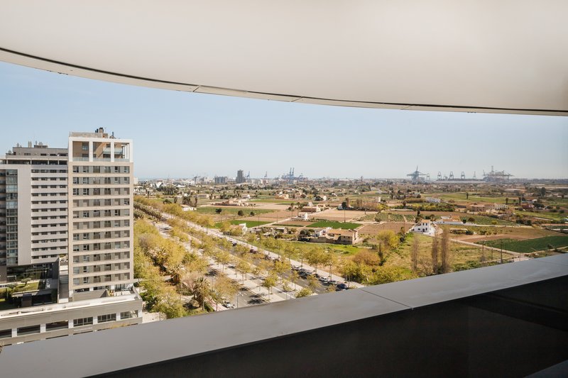 View from a curved balcony across agricultural fields and tree-lined roads toward distant port infrastructure