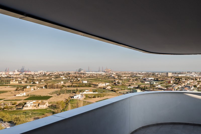 Curved balcony edge beneath dark overhang overlooking rural landscape with industrial skyline in the distance