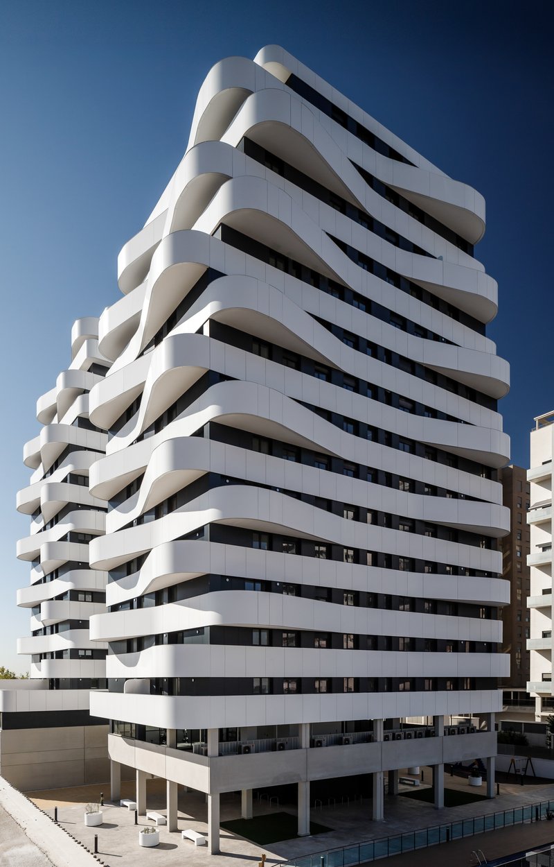 Ground-level view of the tower with stacked undulating balconies on pilotis above a courtyard plaza