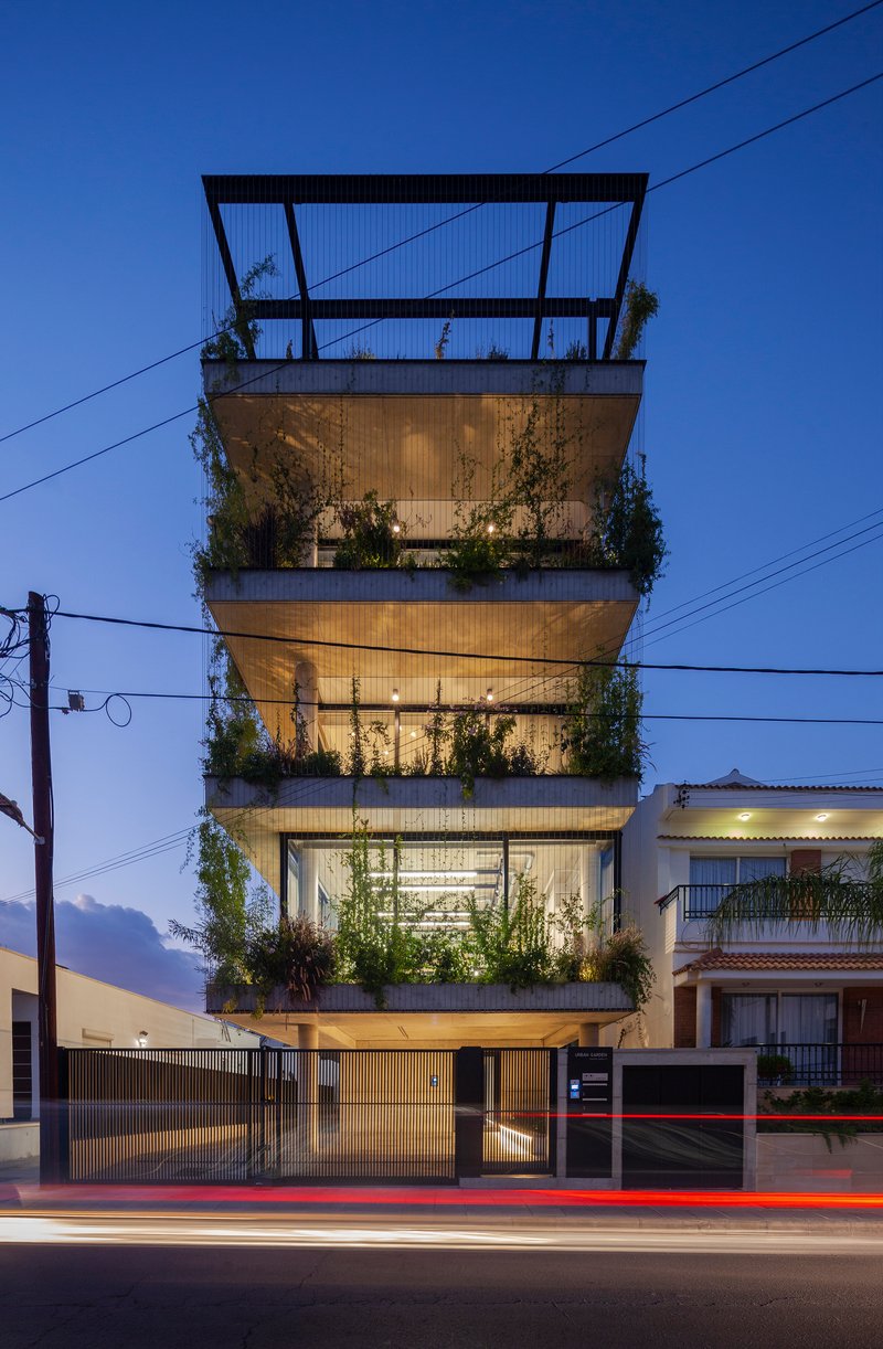 Street view of a four-story concrete facade with planted balconies at dusk with car light trails