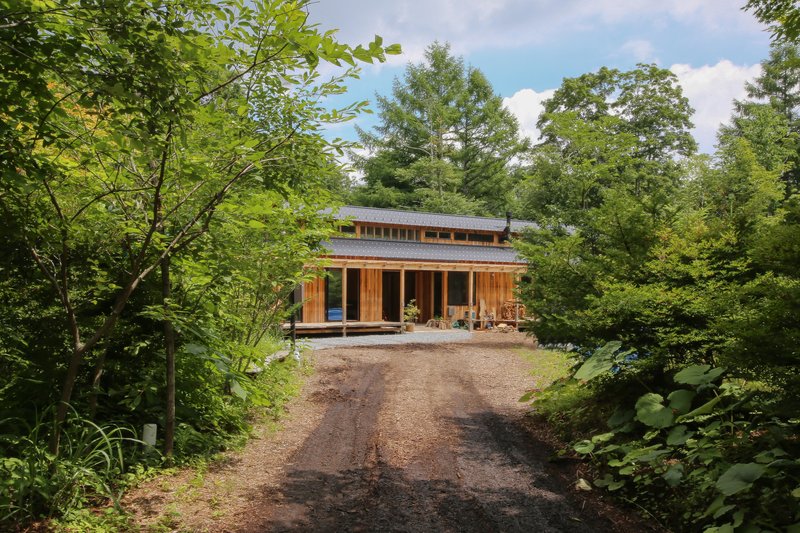 Two-story timber structure with wraparound porch and clerestory band viewed from gravel approach through summer foliage