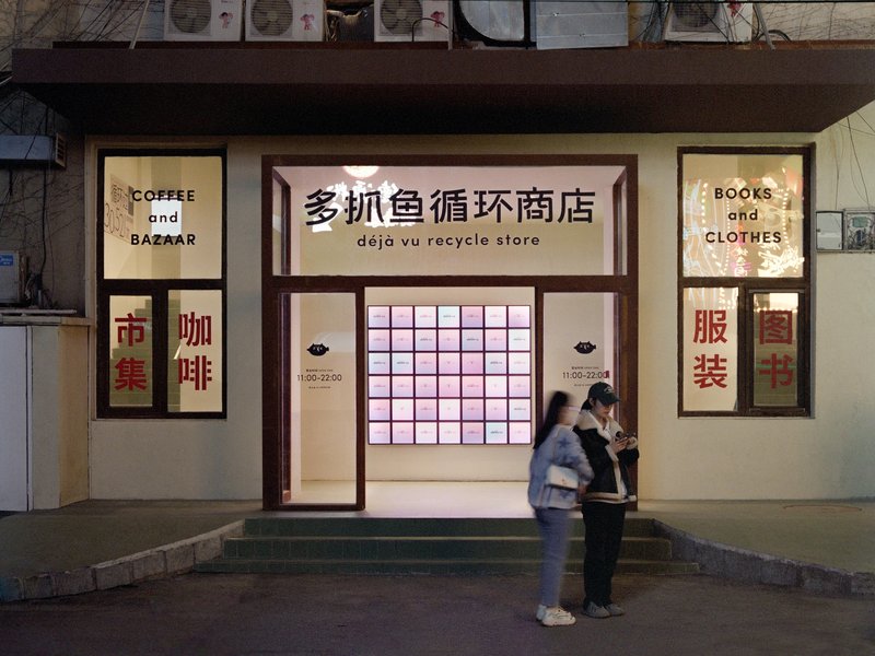Street facade at dusk with dark timber frames, backlit grid panel, and two figures standing outside the entrance
