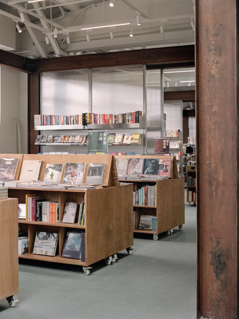 Wheeled plywood display units holding books in front of translucent wall panels and metal shelves beyond timber column