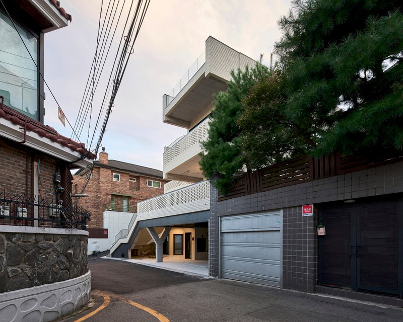 Street view showing stacked volumes with perforated screens and cantilevered terraces beside power lines and pine tree
