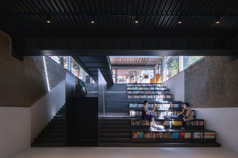 Stepped seating staircase integrated with bookshelves where two readers sit beneath a corrugated metal ceiling