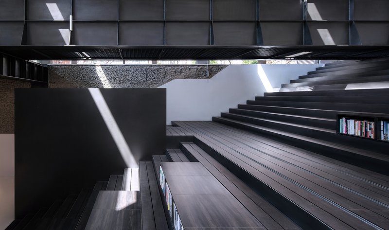 Looking up through the central atrium at black timber stair treads bridging between stone walls with sunlight streaming down
