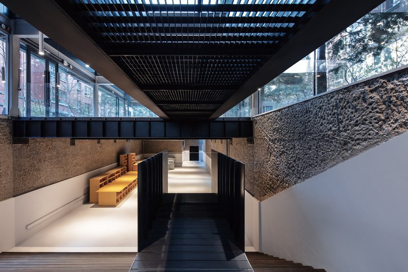 Interior passage showing textured stone walls, black timber overhead beams, and clerestory windows framing adjacent buildings