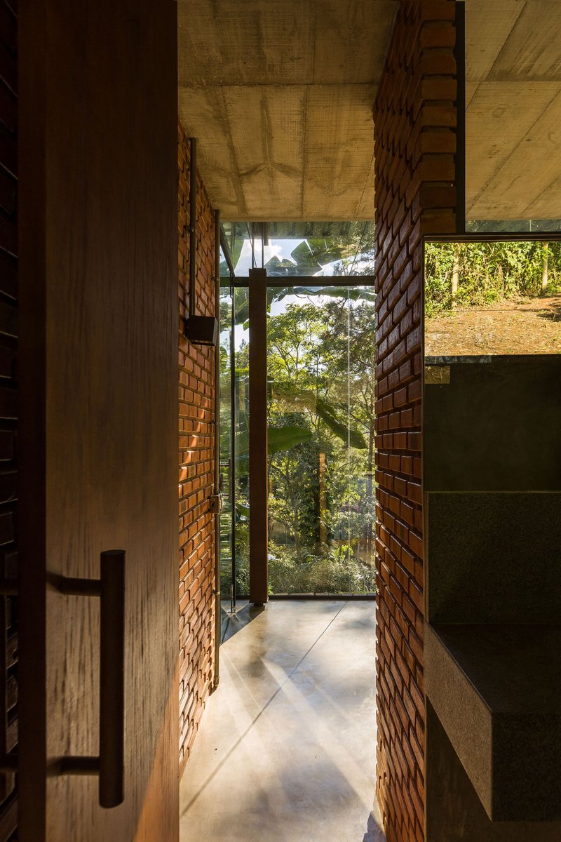 Corridor with perforated brick walls and concrete floor leading to a glass wall framing forest views