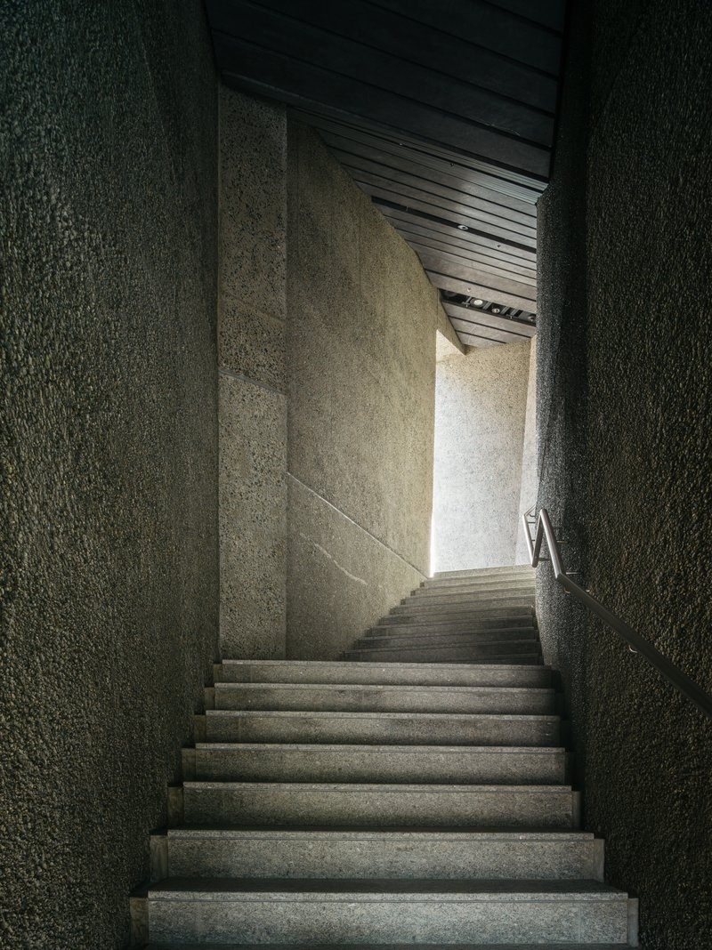 Narrow concrete staircase ascending between textured aggregate walls toward a bright opening above