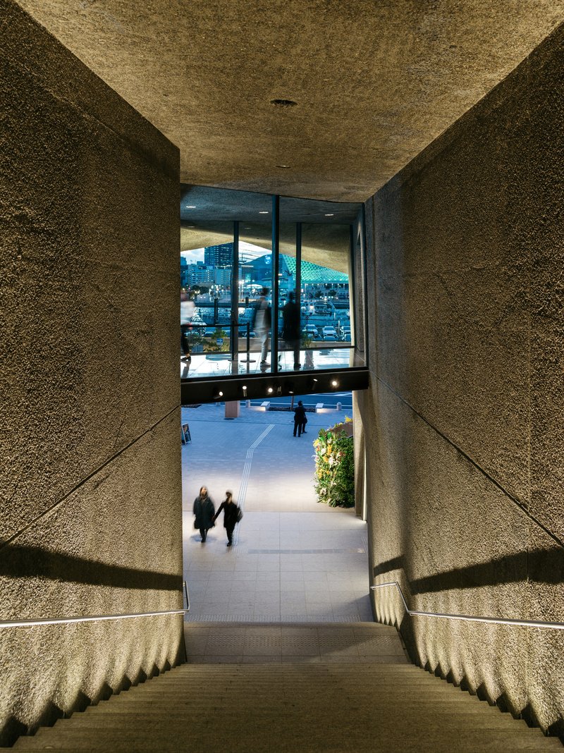 View down the stepped concrete passage with textured walls framing visitors walking toward the plaza at dusk