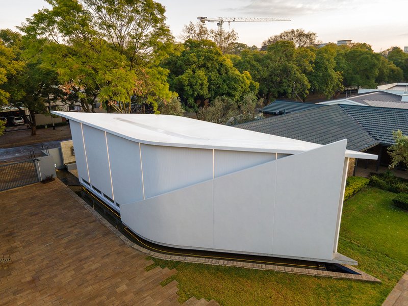 Aerial view of the white curved volume with sloping roof set among mature trees at dusk