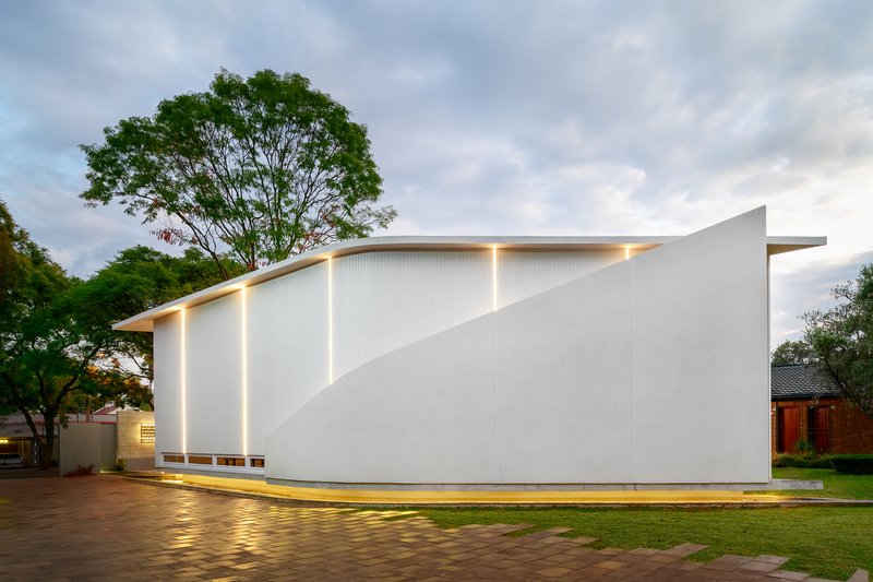 White corrugated metal facade with angled roofline and vertical light slots at twilight beside mature trees