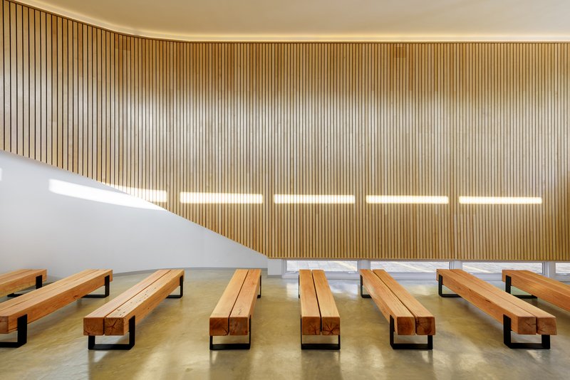 Vertical timber slat wall with five horizontal light strips above rows of wooden pews on concrete floor