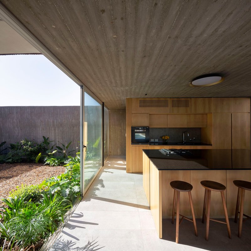 Kitchen with timber cabinetry and board-formed concrete ceiling overlooking planted courtyard through glazed wall