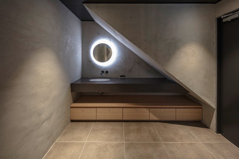 Bathroom vanity tucked beneath a sloped ceiling with circular illuminated mirror at night
