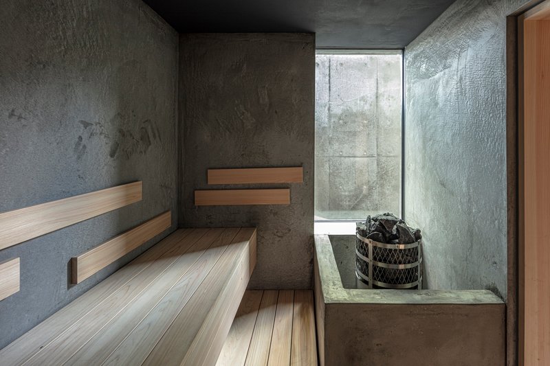 Sauna interior with tiered timber benches and stone heater beside a glass slot window