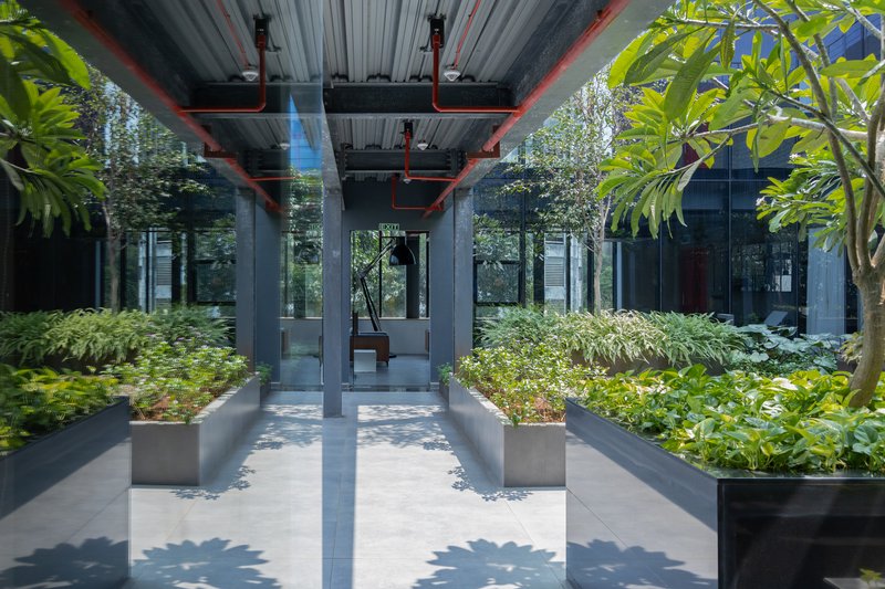 Covered walkway with exposed corrugated ceiling and red structural supports framing planted beds and trees in dappled sunlight