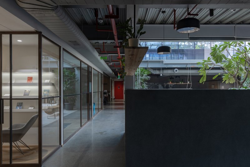 Office corridor with exposed ceiling ducts, glazed meeting rooms, and potted plants along the passage