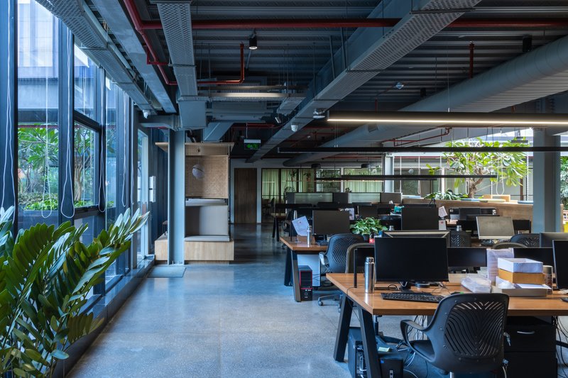 Open office floor with workstations, exposed mechanical systems overhead, and floor-to-ceiling windows admitting daylight