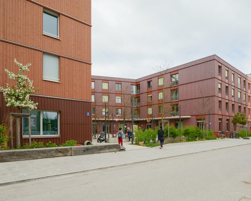 Red corrugated metal facade with residents walking along a paved courtyard under an overcast sky