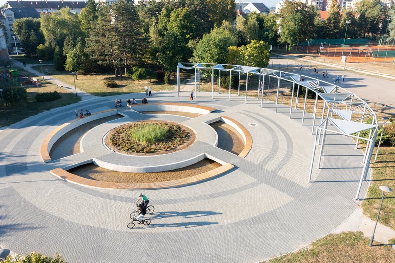 Overhead view of the circular plaza with concentric seating rings and a child cycling across the paved surface