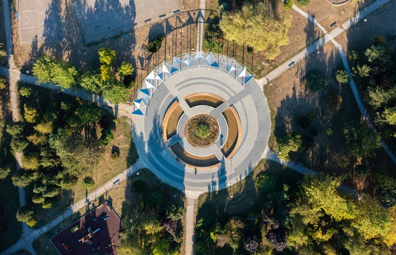 Drone view of the circular pavilion with blue canopy panels nestled among trees casting long shadows
