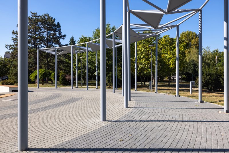 Steel column pergola with triangular shade panels over concentric circular paving under clear blue sky