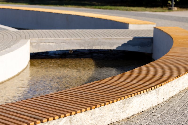 Slatted timber bench curving along the edge of a shallow concrete water feature in afternoon sun