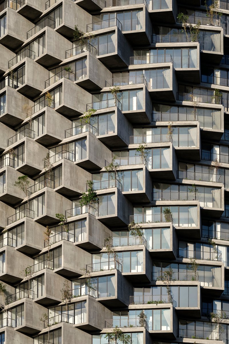 Concrete facade with staggered balconies and floor-to-ceiling glazing planted with young trees in bright sunlight