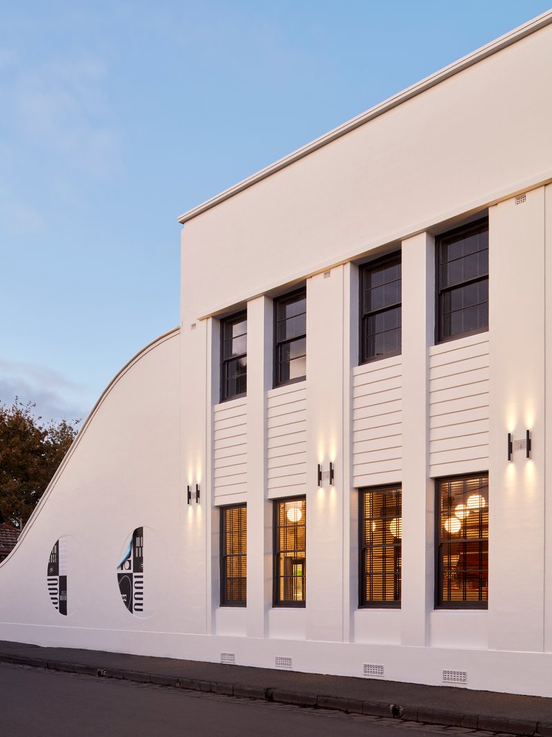White rendered facade with vertical pilasters and clerestory windows illuminated at dusk from the street