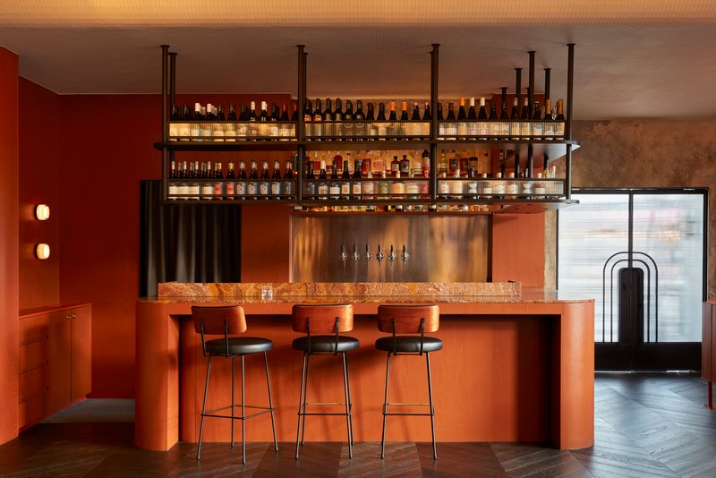 Bar counter with terracotta stone top, suspended shelving, and three bar stools beneath backlit bottles