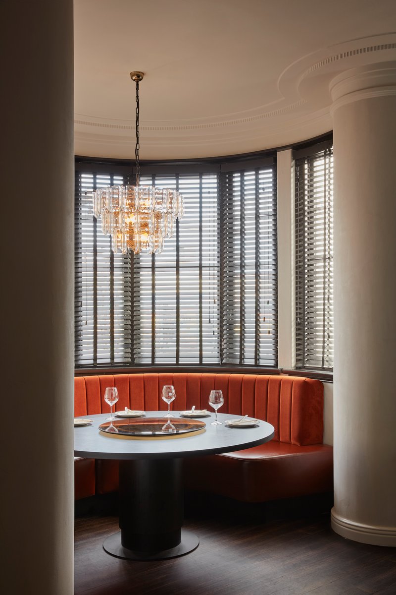 Curved booth with coral banquette, crystal chandelier, and tall windows with venetian blinds at dusk