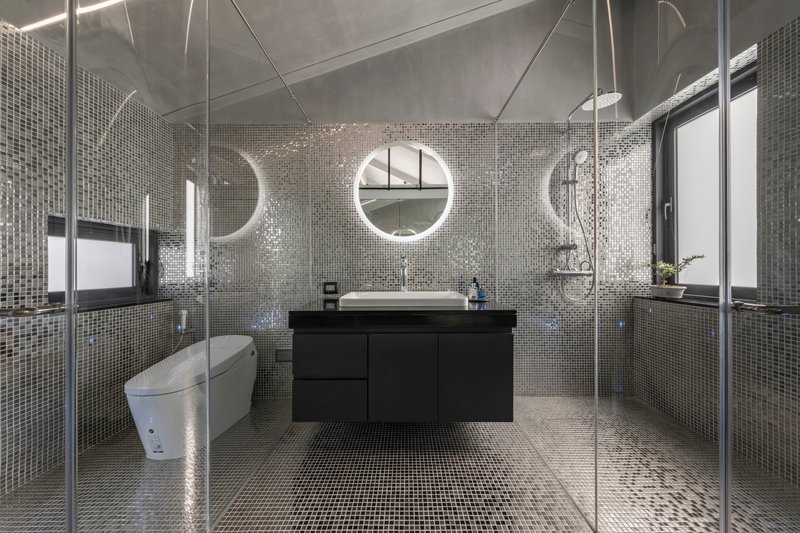 Bathroom clad in reflective silver mosaic tile with glass shower enclosure and round backlit mirrors