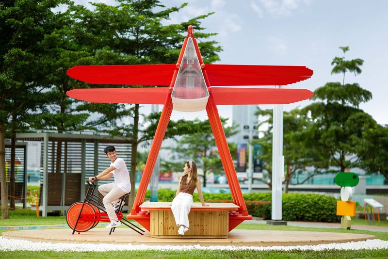 Pavilion with red triangular frame and horizontal canopy sheltering timber seating and two visitors on a sunny day