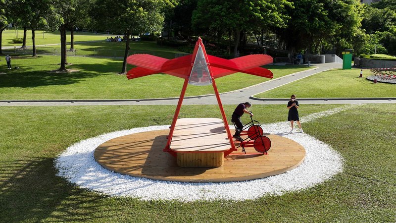 Red steel pavilion with timber deck and white gravel ring in a sunlit park lawn