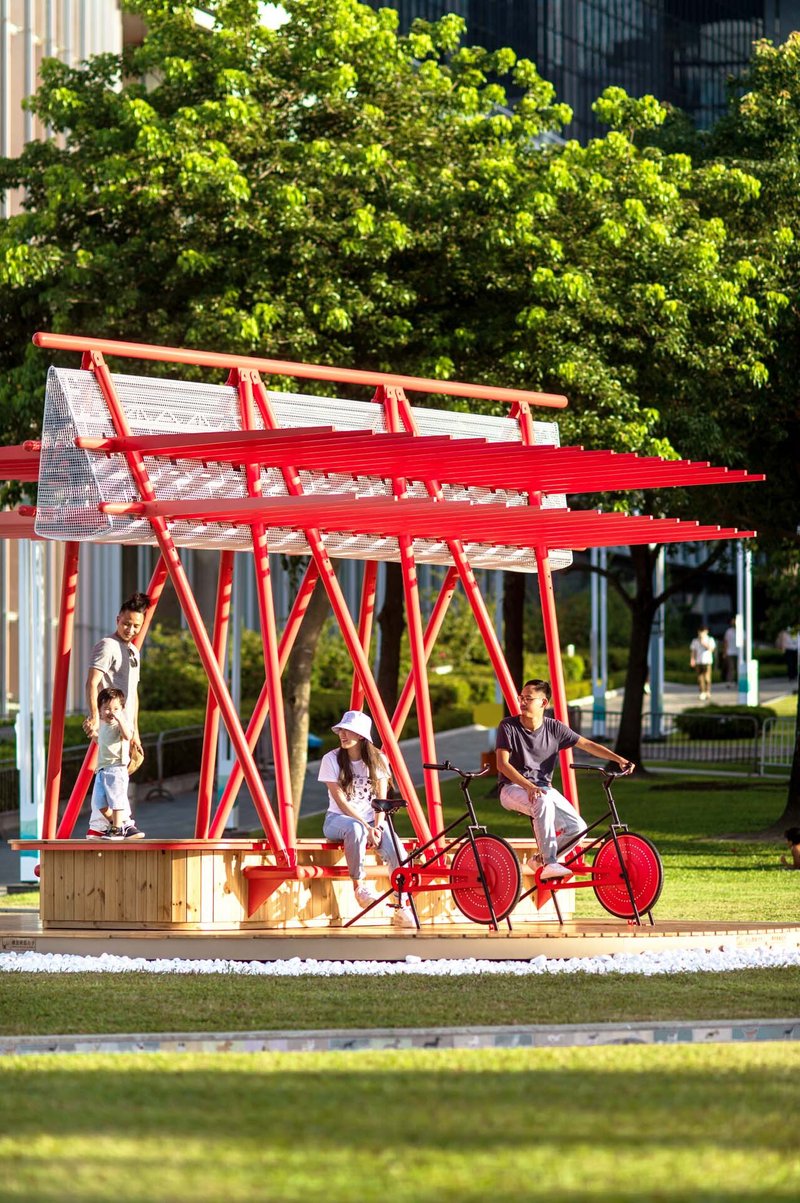 Red steel canopy sheltering a wooden seating platform with visitors on a bicycle installation