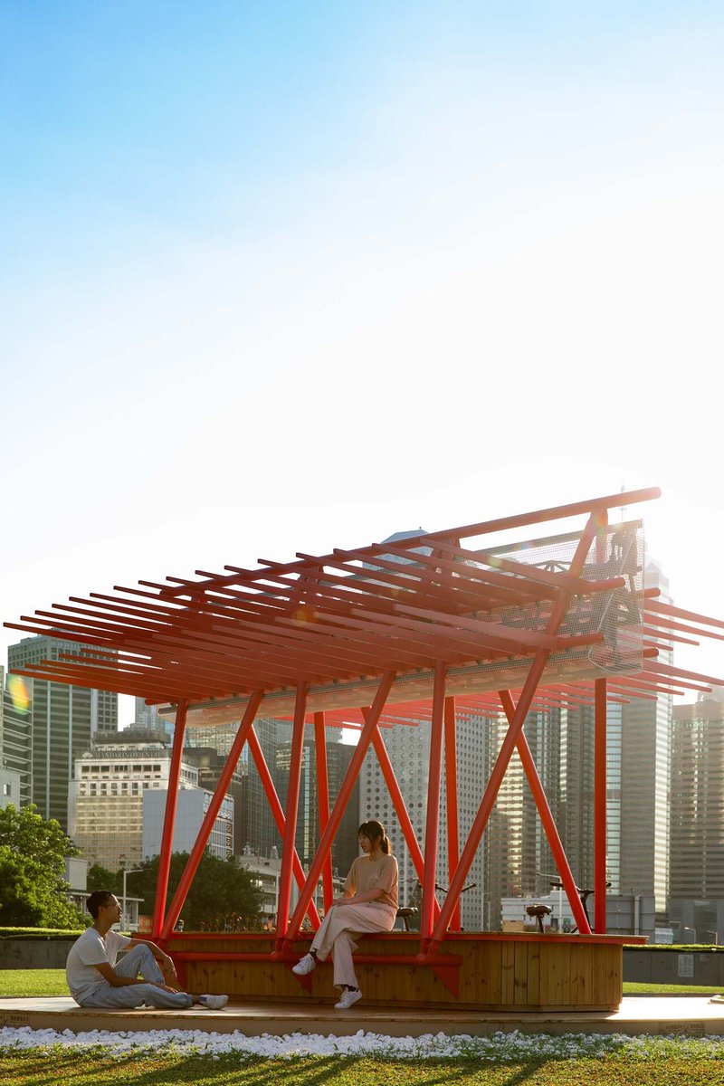 Layered red steel slat canopy above timber bench seating with urban skyline at dusk