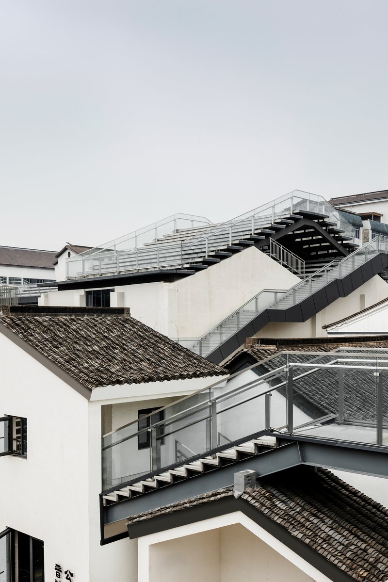 Layered steel staircases zigzagging between white plastered walls and grey-tiled roofs under overcast sky