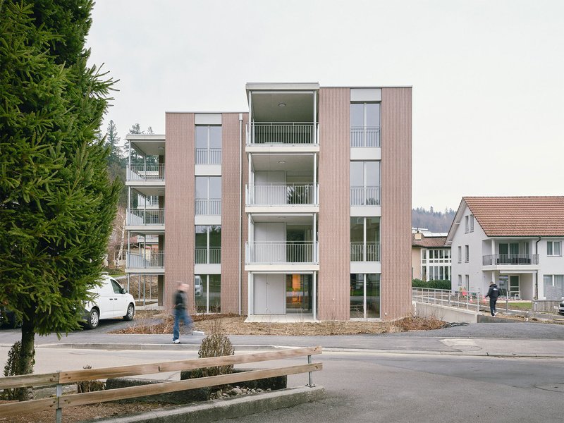 Street view of the four-storey apartment block with ribbed facade and white balconies as a figure walks past